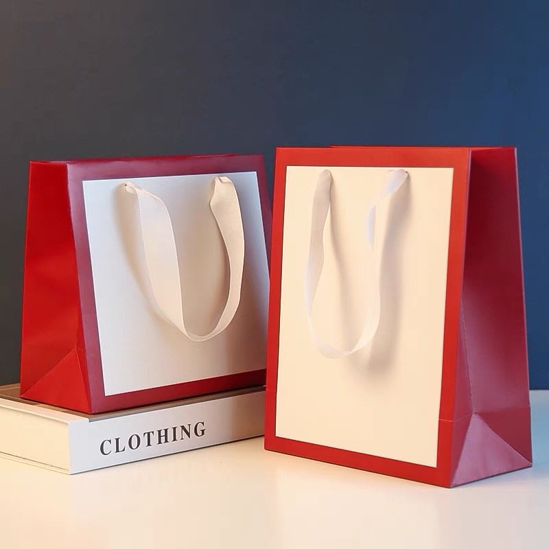 Two red and white paper gift bags with white ribbon handles, placed on a book titled "Clothing."