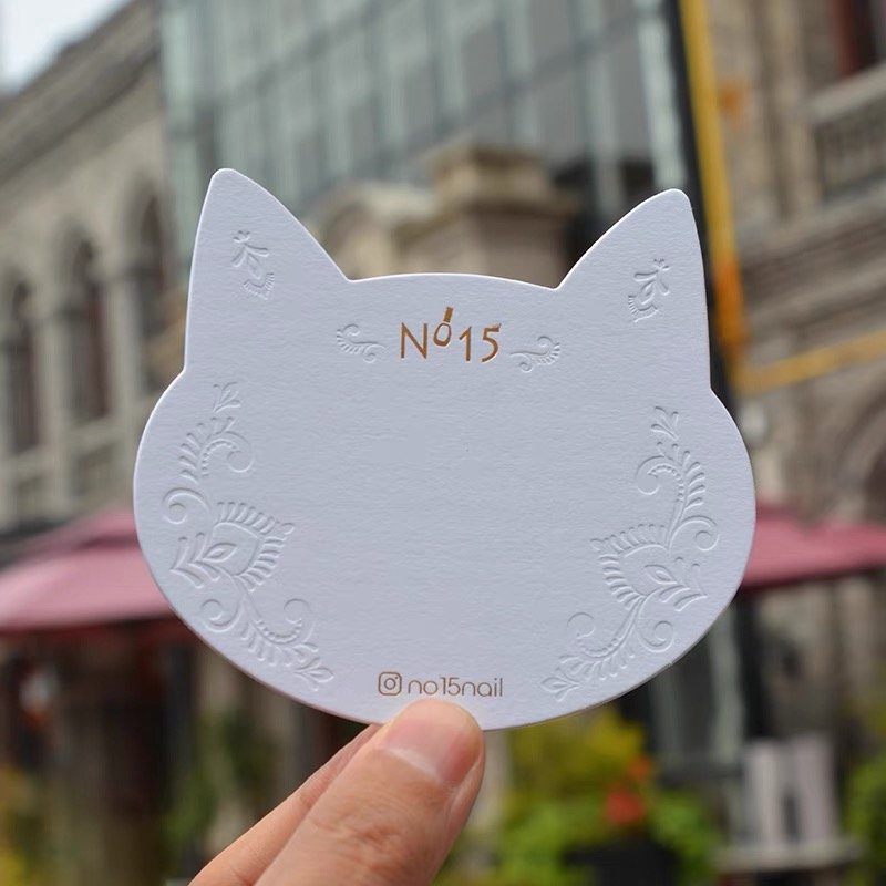 A white, cat-shaped embossed card held outdoors in front of a blurred building.