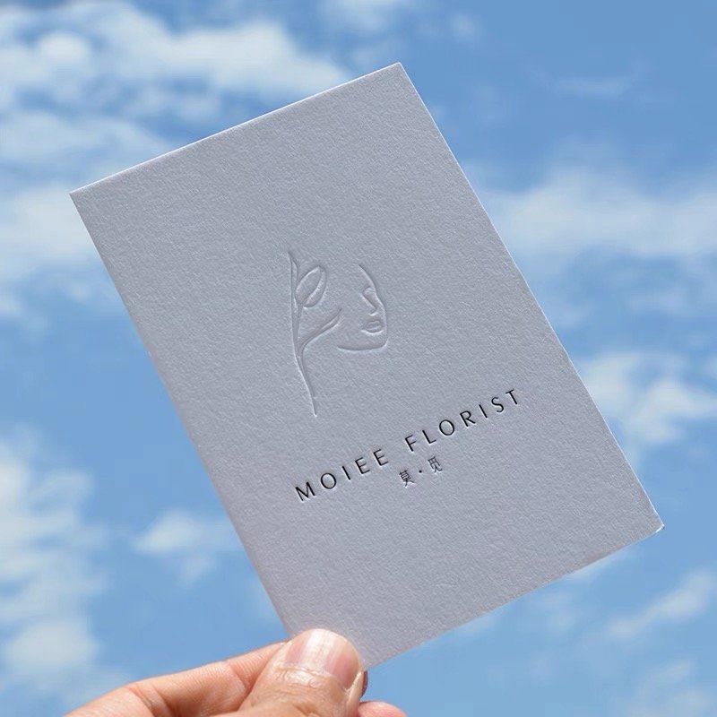White embossed florist card with a logo, held against a blue sky.