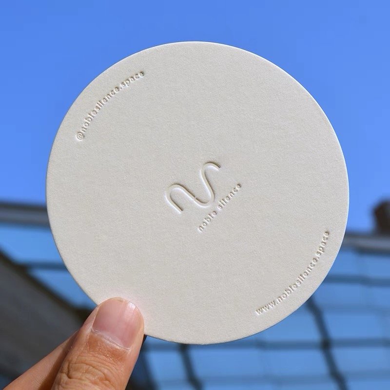 Circular embossed card with "noble silence" logo, held against a clear blue sky.
